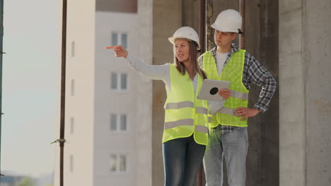 Construction-worker-man-and-architect-woman-in-a-helmet-discuss-the-plan-of-construction-of-house-tell-each-other-about-the-design-holding-a-tablet-look-at-the-drawings-background-of-sun-rays.